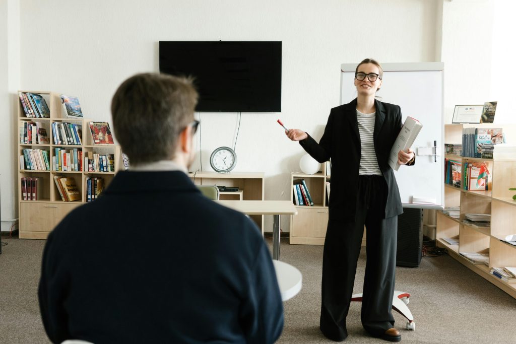 A teacher in a black blazer conducts a session with a student in a library setting.