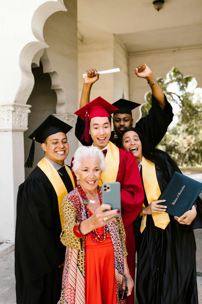 Group of multiracial graduates celebrating with an elderly woman, taking a selfie outdoors.